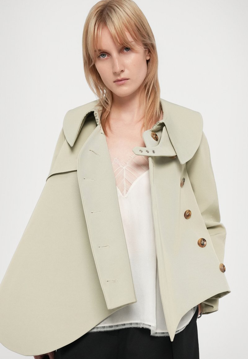 Veste structurée vert clair avec un large col, ornée de gros boutons et d'un design unique en forme de cape, portée par-dessus un haut blanc.