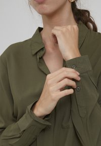 Olivgrön skjorta med krage, knäppta manschetter och bröstficka, tillverkad av ett slätt tyg. Skjortan har en avslappnad passform och långa ärmar.