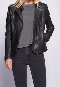 Žena v černé kožené bundě, šedém dlouhém rukávu a tmavě šedých džínách, s jednou rukou v kapse bundy na neutrálním pozadí.