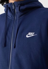 Zbliżenie na osobę noszącą granatową bluzę z kapturem Nike na zamek błyskawiczny z regulowanym kapturem oraz białym haftowanym logo na piersi.