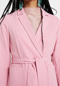 Femme portant un blazer rose ceinturé sur un top côtelé rose clair, avec de longues tresses et de grandes boucles d'oreilles ovales vertes.
