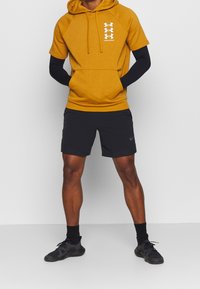 Felpa gialla a maniche corte con cappuccio e logo, abbinata a pantaloni sportivi neri e scarpe da ginnastica nere, che mostra uno stile aderente.