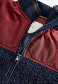 Giacca in pile con corpo testurizzato blu scuro, maniche lisce color bordeaux e colletto a coste navy; include una chiusura con zip e etichetta del marchio.