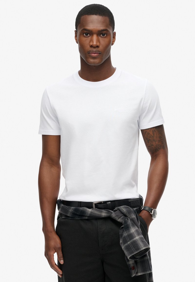 Hvid bomulds t-shirt med rund halsudskæring. Modellen bærer sorte bukser og en gråternet skjorte bundet om livet. Simpelt design, ingen synlige logoer.