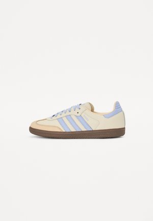 Sneaker basso con tomaia beige, strisce e lacci blu chiaro, e suola marrone texturizzata, mostrata in profilo laterale su sfondo bianco.