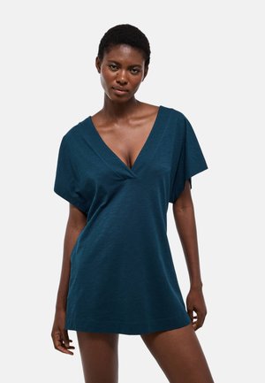 Femme aux cheveux courts portant une robe ample bleu sarcelle foncé à manches courtes et col en V profond, debout devant un fond clair uni.
