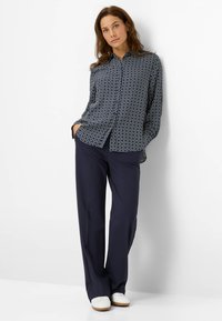 Vrouw in een marineblauwe geometrisch patroon lange mouwen shirt, marineblauwe brede broek en witte schoenen staat met handen in de zakken tegen een witte muur.