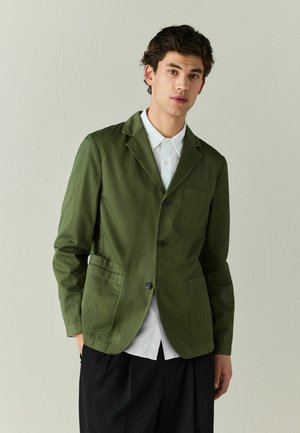 Junger Mann trägt einen grünen Blazer über einem weißen Hemd und schwarzen Hosen, steht vor einem einfarbigen hellgrauen Hintergrund.