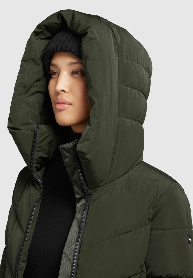 Dunkelgrüne Pufferjacke mit einer großen, strukturierten Kapuze, Reißverschluss und gestepptem Material. Verfügt über ein schwarzes Futter und eine Logostickerei.