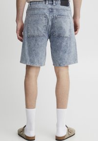 Denim shorts in light blue with a faded finish, featuring two back pockets and a straight cut. Worn with white socks and brown sandals.
