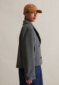 Grijze wollen jas met een dubbelbreasted ontwerp, grote revers en decoratieve knopen. Gekleed met een tan baseballpet en denim onderkleding.