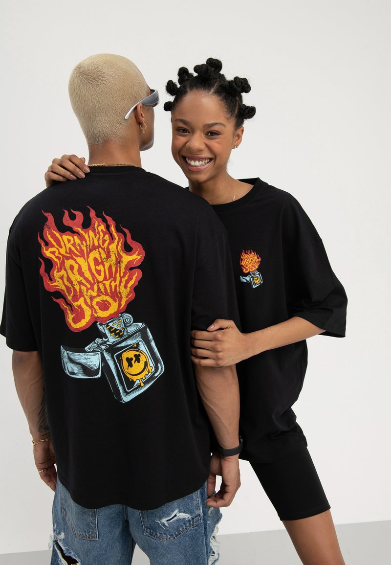 Two people wearing black graphic tees, one facing back showing a flaming lighter design, the other smiling with hand on the other's arm.