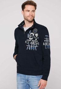 Sudadera azul marino con cremallera de un cuarto, con impresiones gráficas blancas y un parche en el pecho. Material de algodón suave, bolsillo frontal.