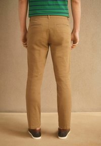 Homme debout de dos, portant un pantalon beige, une chemise rayée verte et bleue, et des baskets noires à semelles blanches sur fond beige.