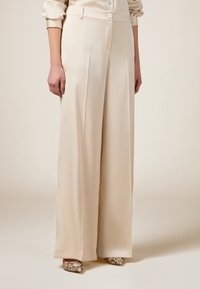 Beige Satin-Hosen mit weitem Bein, seitlichen Einsätzen, Knopfverschluss und glatter Textur, kombiniert mit gemusterten Schuhen.