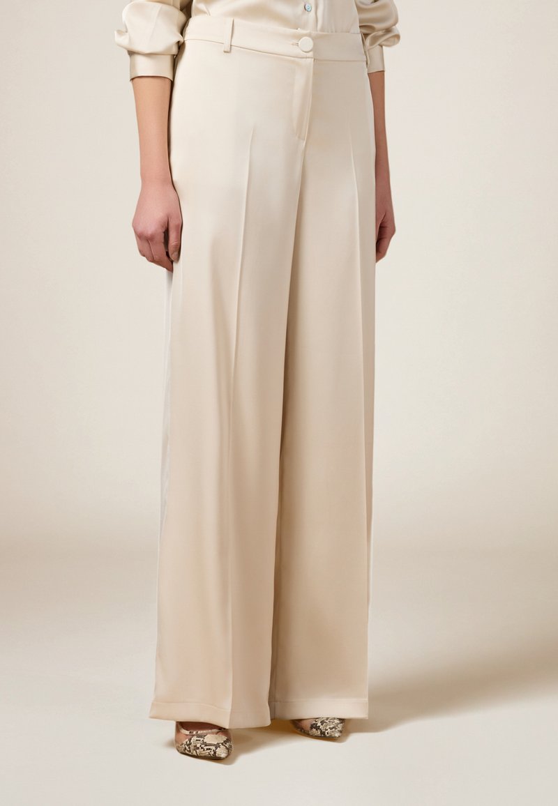 Beige Satin-Hosen mit weitem Bein, seitlichen Einsätzen, Knopfverschluss und glatter Textur, kombiniert mit gemusterten Schuhen.