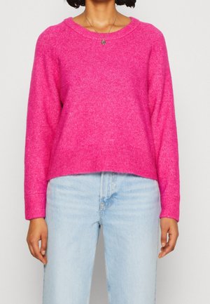 Žena v jasně růžovém svetru s dlouhými rukávy a světlých modrých džínách s vysokým pasem, s viditelným malým kulatým náhrdelníkem.