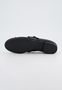 Zapato negro de cuero con suela plana, que cuenta con un acento de hebilla. La suela es lisa con un logo grabado, y el diseño general es estilizado.