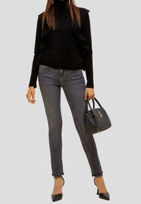 Suéter negro con hombros con volantes, combinado con jeans skinny oscuros. La modelo sostiene un pequeño bolso negro y lleva puestos unos tacones negros de punta.