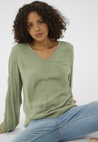 Blouse vert clair à manches longues avec un col en V et un détail de couture subtil au centre. Portée avec un jean bleu, à la coupe décontractée.
