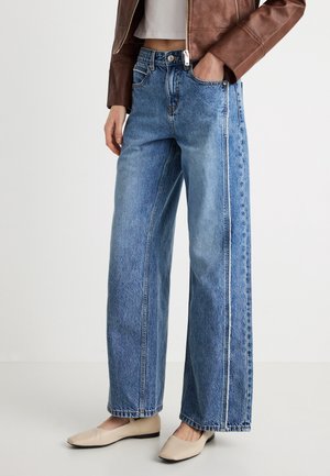 Világoskék, bő szárú farmernadrág, magas derékkal, oldalzsebekkel és kontrasztos varrással. Bézs lapos talpú cipővel párosítva.