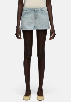 Person iført lyseblå og hvide stribede denimshorts, cremefarvede flade sko og en indstukket lys top, står med afslappede arme.