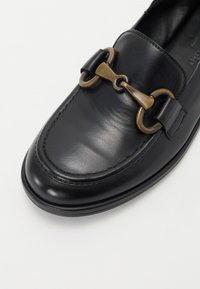 Zwarte leren instappers hebben een ronde neus, zichtbare stiksels en een metallic hardware accent met een gespdetail.