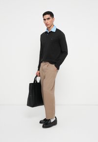 Mladý muž stojí proti bielej stene, oblečený v čiernej blúzke s dlhým rukávom a modrým golierom, béžových nohaviciach, čiernych mokasínach, bielych ponožkách a drží čiernu tašku.