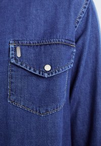 Denim särgil on rinna tasku hõbedase nupu ja sinise niidi, millel on kerge tekstuur ja kontrastne kollane õmblus.