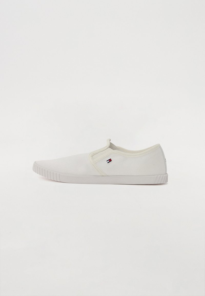 Fehér vászon slip-on cipő texturált talppal, az oldalán egy kis színes logóval és ívelt kialakítással az orr résznél.
