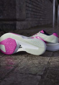 Hvide og pink Adidas Adizero løbesko placeret på en stensat flisegang, hvor den ene sko viser sålen og mærket.