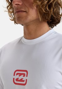 Biała koszulka sportowa z czerwoną falą logo na klatce piersiowej. Posiada okrągły dekolt i gładki, lekki materiał.
