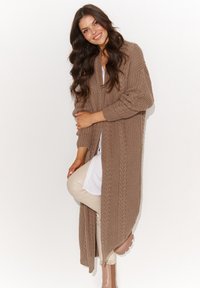 Długi kardigan z otwartym przodem, wykonany z teksturowanego, jasnobrązowego dzianiny. Posiada ściągacze przy mankietach oraz wzór w warkocze. Noszony na białej bluzce i jasnych spodniach.