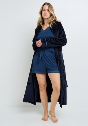 Robe en velours bleu marine porté sur un ensemble de débardeur et de shorts assortis. La robe possède des manches longues et une taille à nouer. Détails en dentelle sur les shorts.