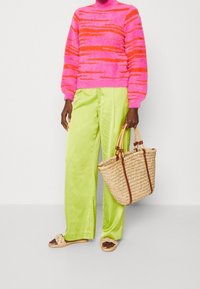 Maglione fuzzy a righe rosa brillante e arancione, pantaloni a gamba larga in satin verde, sandali intrecciati e borsa in paglia con accenti marroni.