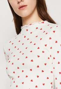 Frau trägt ein weißes, geripptes Top mit kleinem roten Herzmuster und Rüschen am hohen Kragen, ihr Gesicht von den Schultern bis zum Kinn sichtbar.