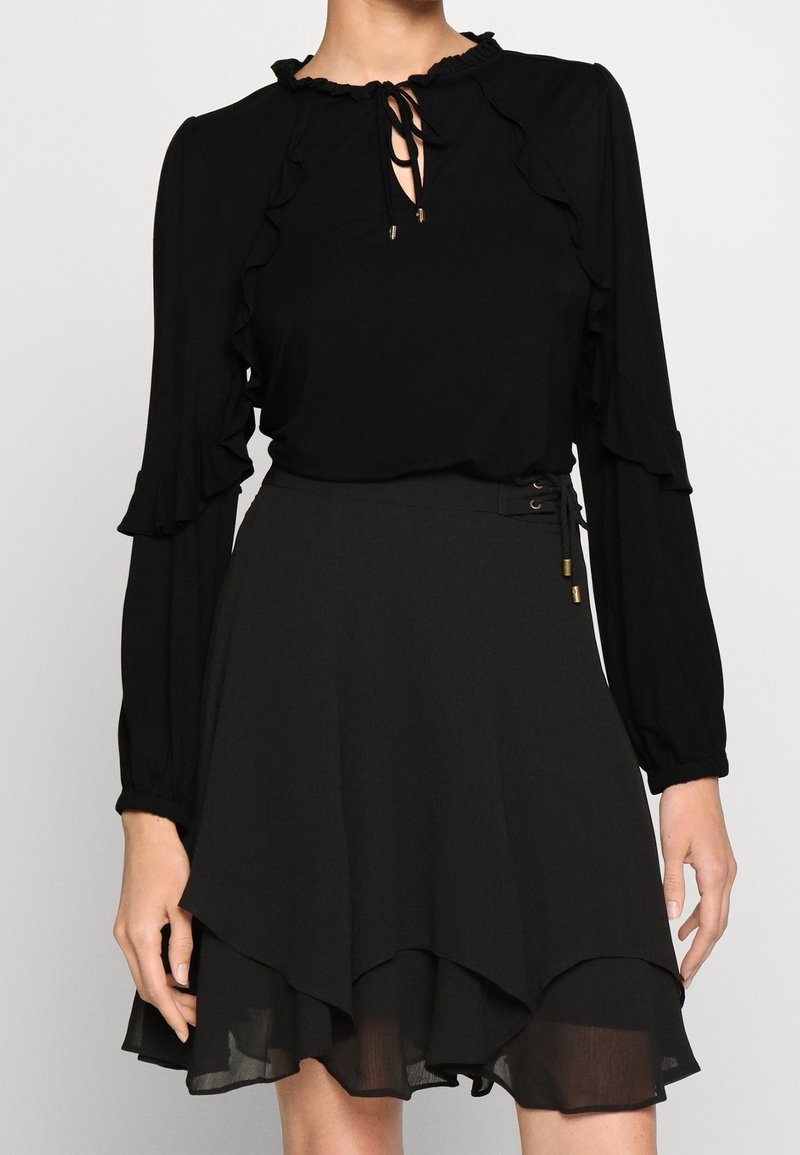 Svart blus med volanger och knytkrage, kombinerad med en matchande utsvängd kjol med lagerkant och dekorativ snörning på sidan.