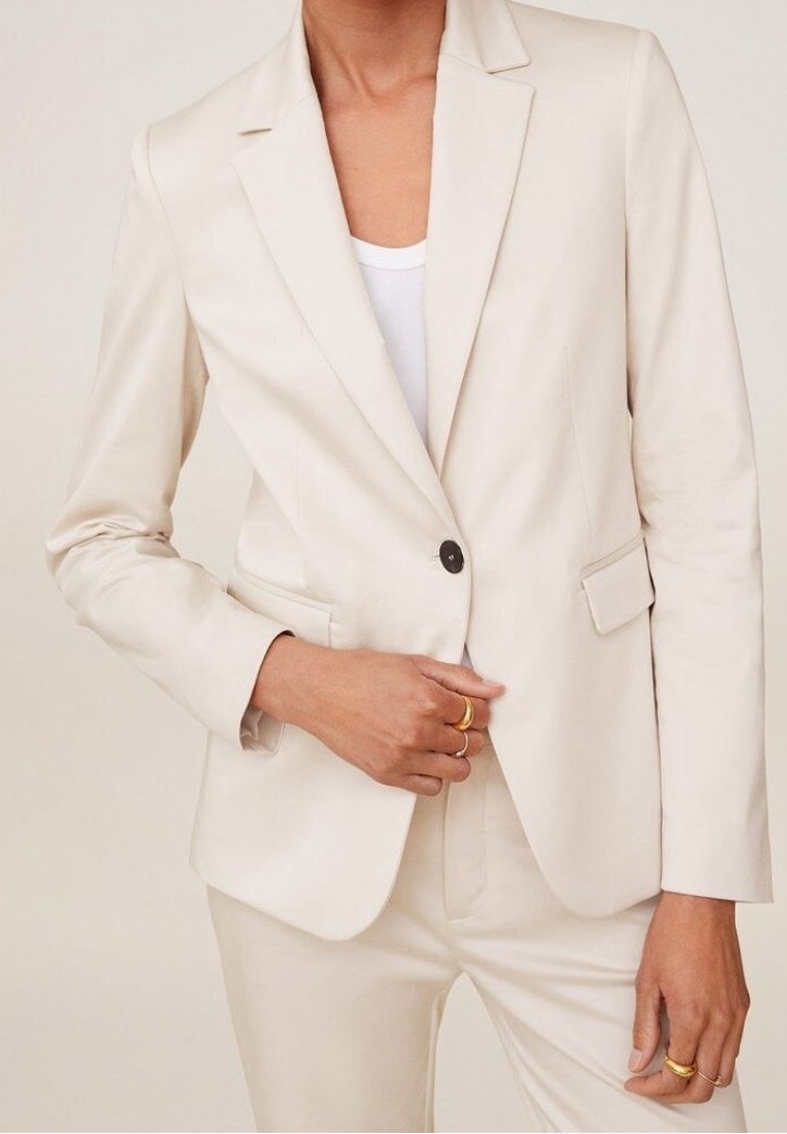 Blazer beige sur mesure avec un seul bouton noir, revers crantés et texture lisse, associé à un pantalon beige assorti.