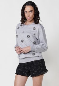 Felpa grigia con decorazioni argentate, scollatura rotonda e maniche lunghe, abbinata a pantaloni corti neri testurizzati con pulsante e orlo sfrangiato.