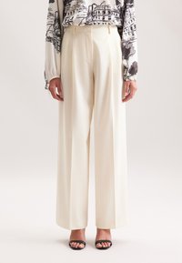 Vrouw in crème kleurige hoge taille, wijdvallende broek, gecombineerd met een gebloemde blouse met lange mouwen en zwarte open-tong hakken.