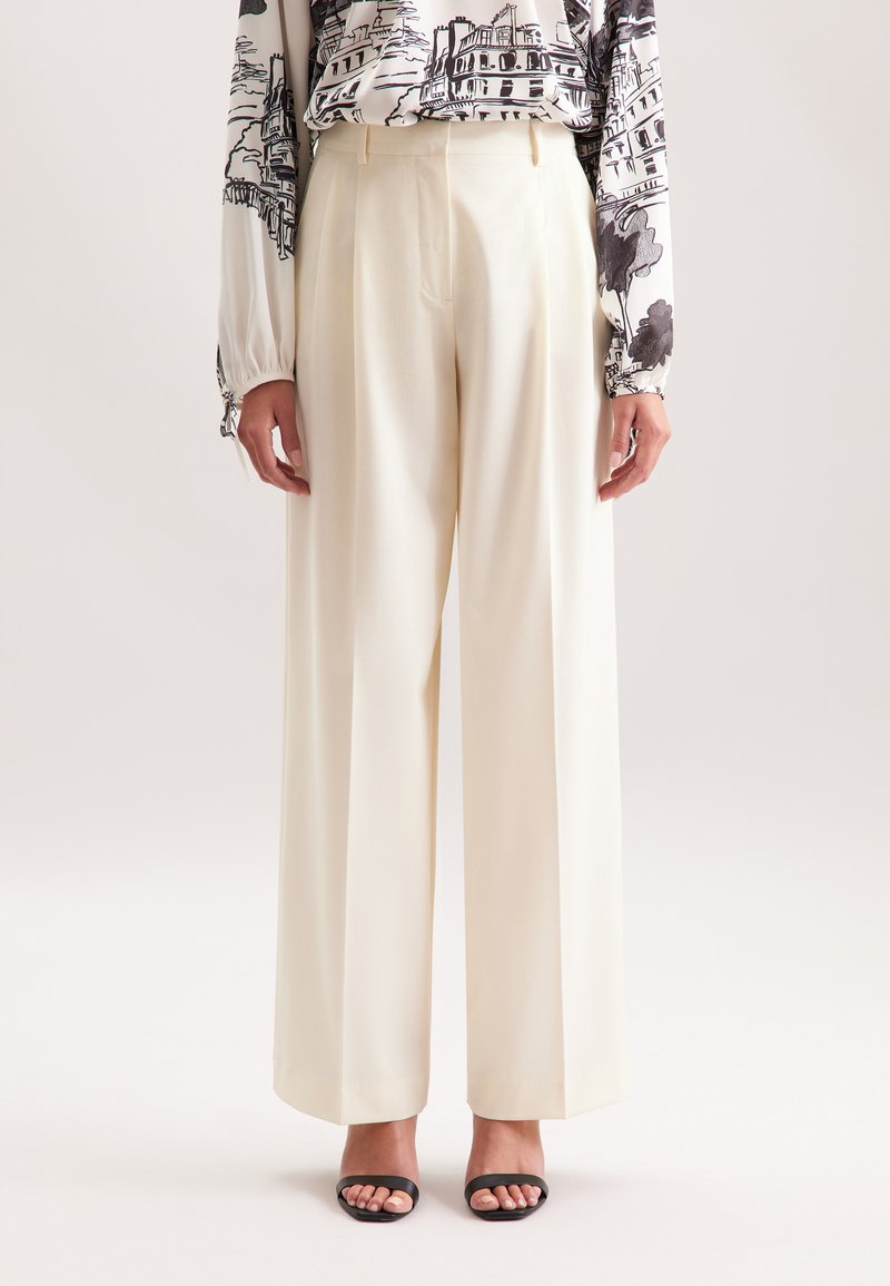 Vrouw in crème kleurige hoge taille, wijdvallende broek, gecombineerd met een gebloemde blouse met lange mouwen en zwarte open-tong hakken.