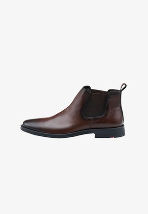 Bottines Chelsea en cuir marron avec panneaux élastiques sur les côtés, bout arrondi et tirant; semelle plate noire avec un accent rouge près du talon.