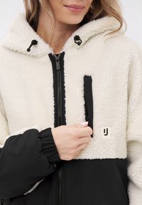 Fleece-lined hooded jacket featuring a cream upper and black lower section, zip closure, and a visible logo on the chest pocket.