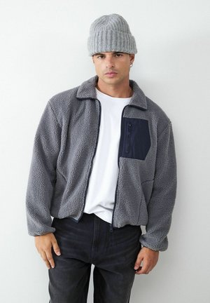 Mladý muž s šedou pletenou čiapkou, šedou fleecovou bundou s čiernym náprsným vreckom, bielym tričkom a čiernymi nohavicami na obyčajnom bielom pozadí.