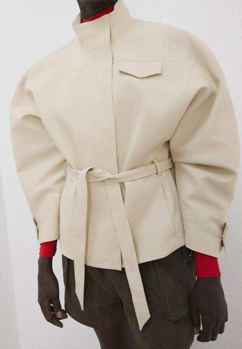 Beige belted jacket with structured shoulders worn over a red long-sleeve top and dark brown pants.