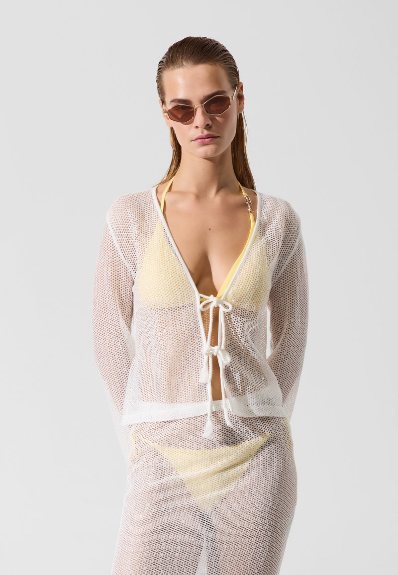 Žena v žltých bikinách so sieťkovaným bielym prehozom a slnečnými okuliarmi, stojaca na svetlom šedom pozadí.