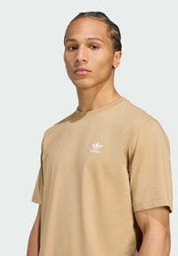 Beige katoenen t-shirt met een ronde hals, korte mouwen, met een klein wit Adidas-logo op de linkerkant van de borst. Glad textuur, ontspannen pasvorm.