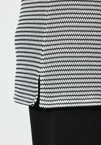 Černobílý vzorovaný top se strukturovaným tkaným designem, rovného střiženého s bočními rozparky, spárovaný s jednoduchým černým spodkem.