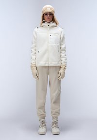 Witte fleece jack met een voorzak, beige broek, beige handschoenen en bijpassende muts. De outfit heeft zachte texturen en een neutraal kleurenpalet.