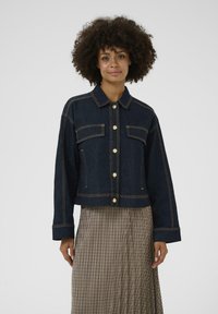 Lühendatud tume teksajakk kuldsete nööpidega, kahekordse rinnataskuga ja silmapaistva kollase õmblusega, kantud ruudulise seeliku peal.